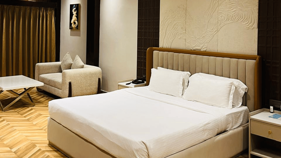 A modern hotel room with a king size bed, beige sofa, white coffee table, wooden herringbone flooring, warm lighting, and a gold and blue abstract artwork on the wall - Silver Sand Beach Resort, Havelock