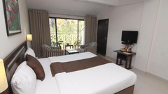 A room interior with a made-up bed featuring white linens and brown accents. A glass door is visible leading to a balcony - Abad Copper Castle, Munnar