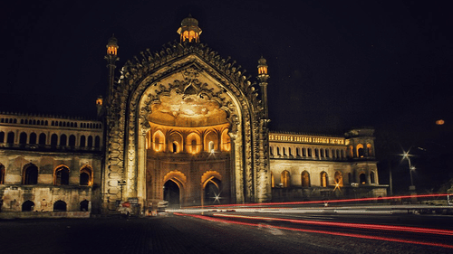A historic, grand arched gateway is illuminated at night, with light trails from cars on the street in front of it.