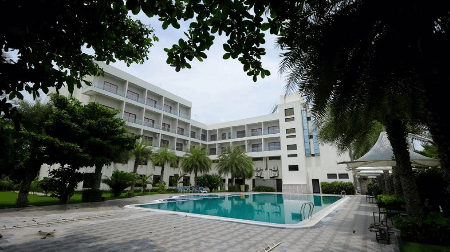 The Arawali Ananta Elite Jaipur hotel building with a large swimming pool and several palm trees.
