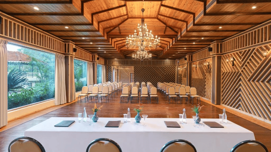 Front view of The Grandeur banquet hall featuring a white table lined with 4 chairs, overlooking the hall with chairs arranged in theatre setting - Golden Tulip Goa Mandrem