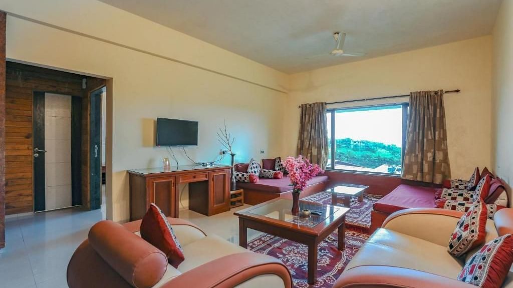 a living room with sofas placed around a table, a mounted television and a large window - Camel Valley Resort, Igatpuri