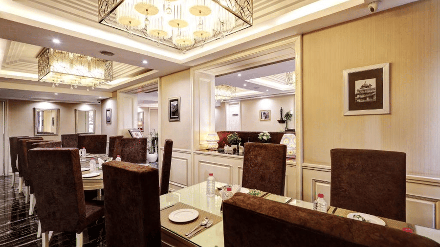 A low-angle perspective of an upscale dining room featuring dark velvet high-back chairs, white panelled walls, and warm square chandeliers - Flute Boutique By Hotel 91, Jaipur