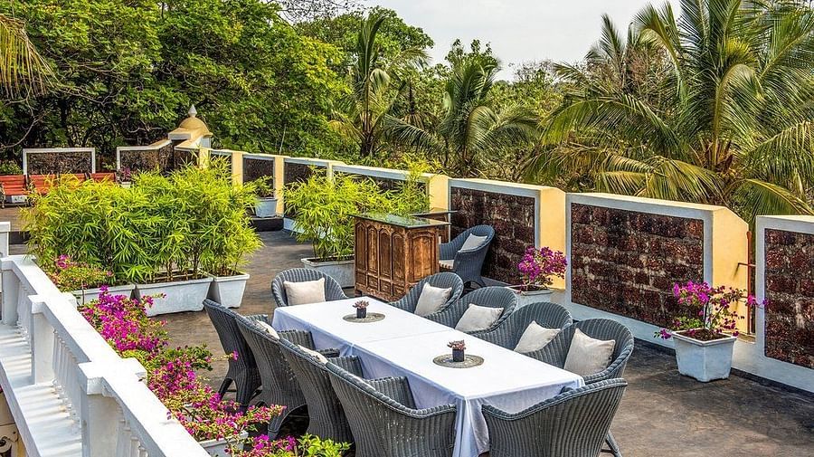 An outdoor dining area on a patio or terrace with a white railing, featuring a long table set with chairs. Lush green plants and trees surround the area at Fort Tiracol, Goa.