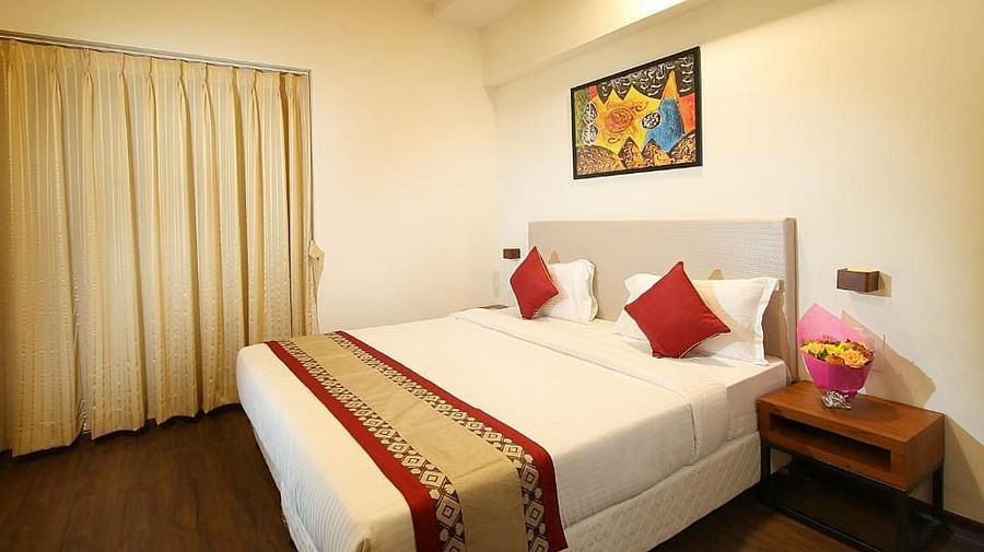 Another bedroom with a double bed adorned with red accents, a wooden side table, a wall-mounted lamp, and a large window with a city view.