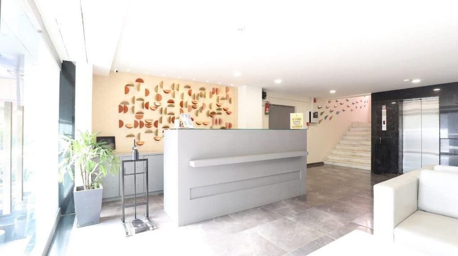 Side view of a brightly-lit reception featuring the front desk at Pravasa Stay, Ahmedabad (Alba Premier), along with an elevator at the far end of the lobby.