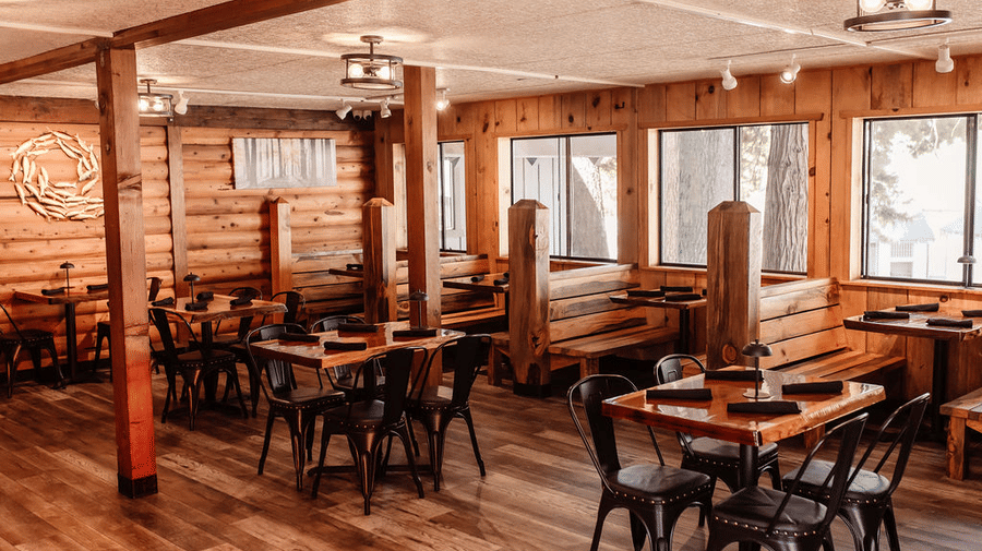 A restaurant with wooden interiors featuring black chairs and tables, wooden pillars and wide windows at Shaver Lake Village Hotel.