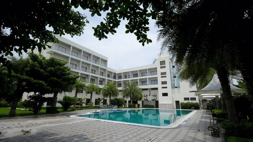 The Arawali Ananta Elite Jaipur hotel building with a large swimming pool and several palm trees.