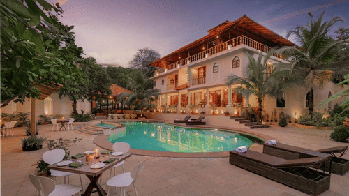 Exterior of Golden Tulip Goa, Mandrem Beach at dusk with a curved pool, lit balconies, palm trees, sun loungers, and a poolside dining table.
