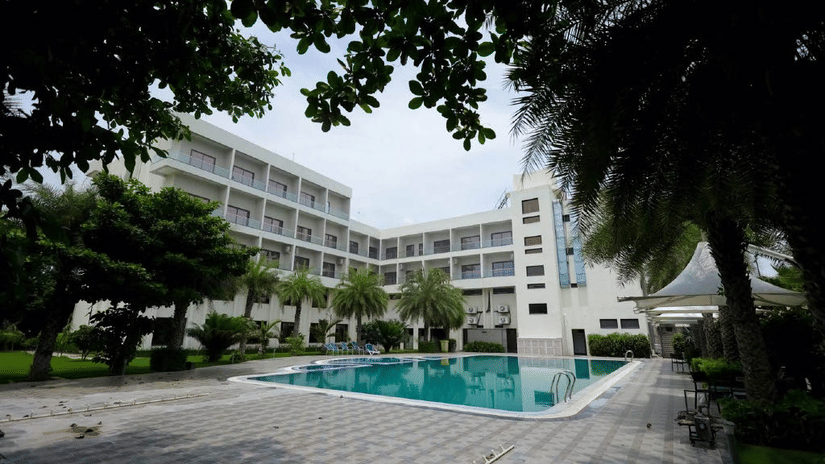 The Arawali Ananta Elite Jaipur hotel building with a large swimming pool and several palm trees.
