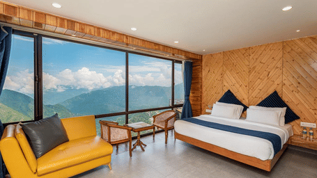 Modern room at MV Acosta, Mussoorie featuring a double bed, yellow sofa, and scenic mountains visible through large windows.