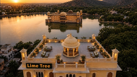 A view from above Lime Tree Hotel & Banquet, Jaipur. The hotel stands next to a lake with a structure in the water and hills in the background.