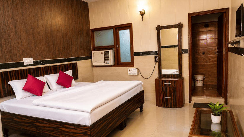 Guest room interior of Hotel Goverdhan, Agra, featuring king size bed, 2 pillows, wooden headboard, dressing table with mirror, window air conditioner, glass table, plant, and door.