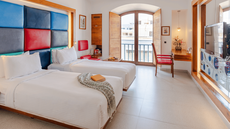 Wide angle view of a room at The Promenade Pondicherry with twin beds, a multicoloured headboard, arched windows, balcony access, and natural light.