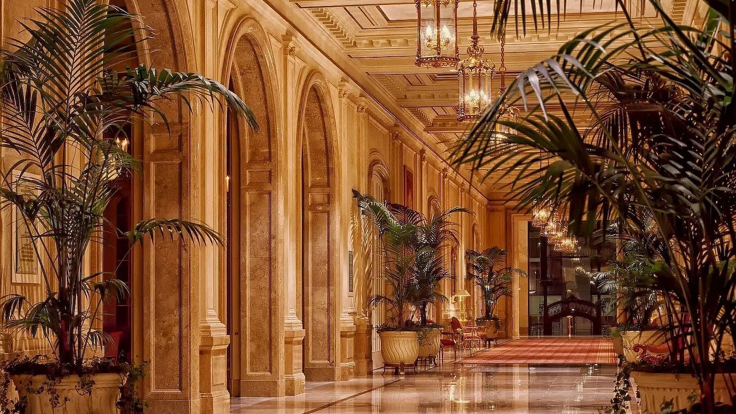  Ornate hallway with arched doorways and potted plants, warm lighting.