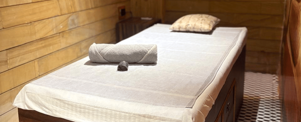 Massage bed with white linen, a rolled towel and a cushion against warm wood panelling in the SKYN Spa therapy room