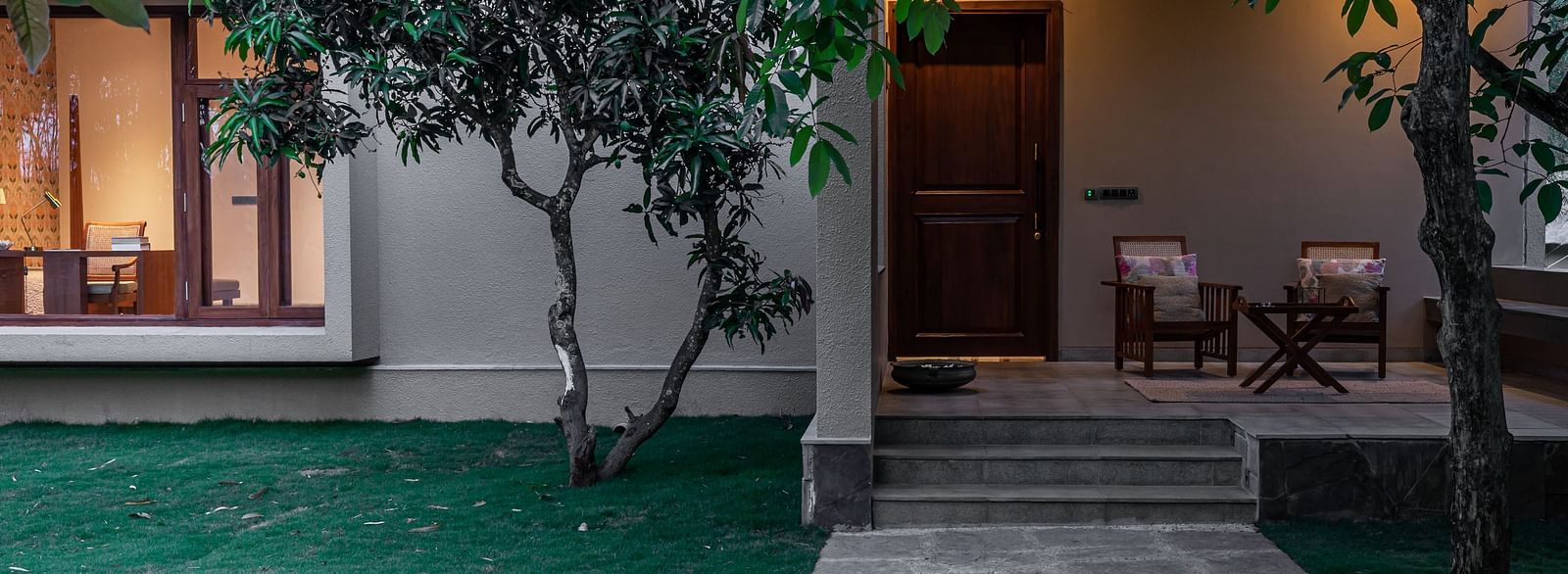 Exterior of a building with lit windows, trees, and a stone pathway at dusk - Shreyas Retreat