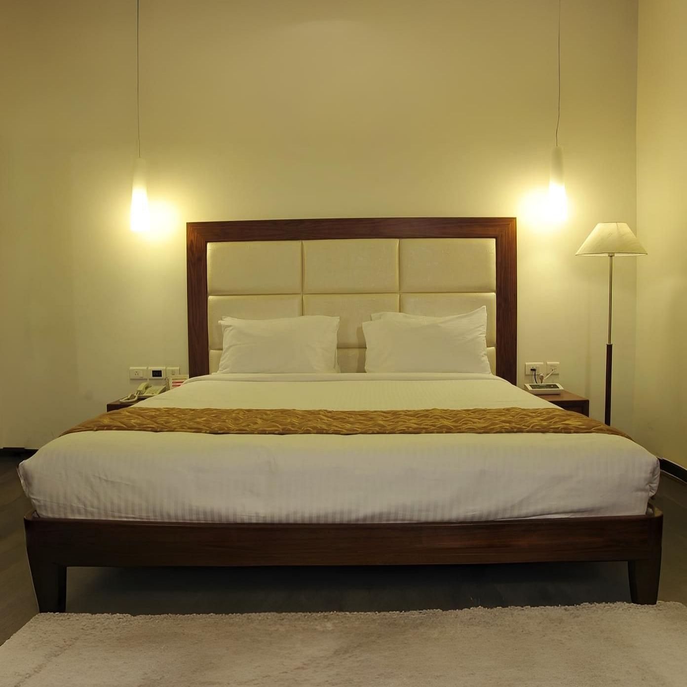 A queen size bed with white linens and a gold runner, two hanging lights, a floor lamp, and a white rug on a wooden floor at Dhanalakshmi Srinivasan Hotels, Perambalur.