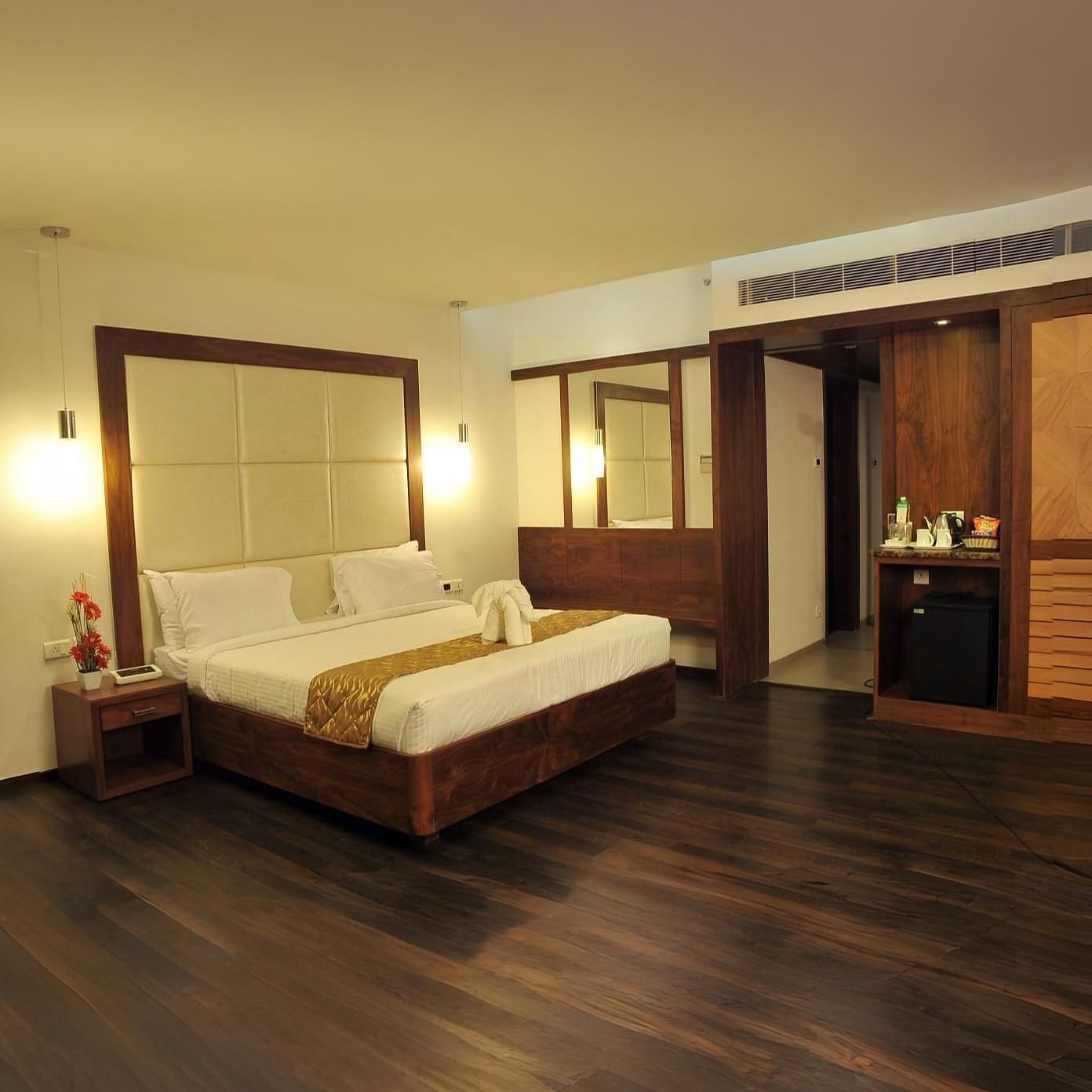 A room with a queen size bed, two side tables, wooden flooring, two mirrors, hanging lights, and a counter with bottles and appliances at Dhanalakshmi Srinivasan Hotels, Perambalur.
