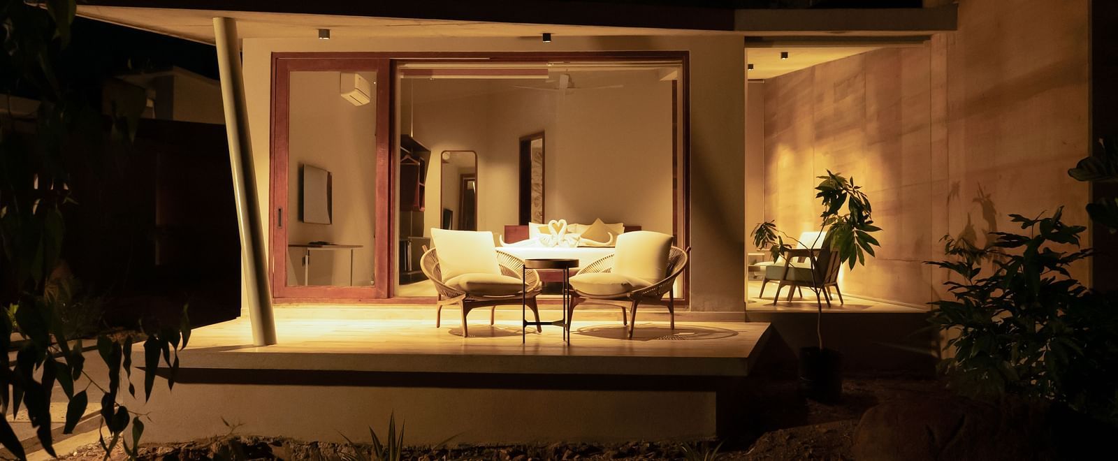A warmly lit interior space with minimalist furniture and a large opening looking out to a dark exterior | Gaia Studio | RD's Nature Retreat, Bangalore