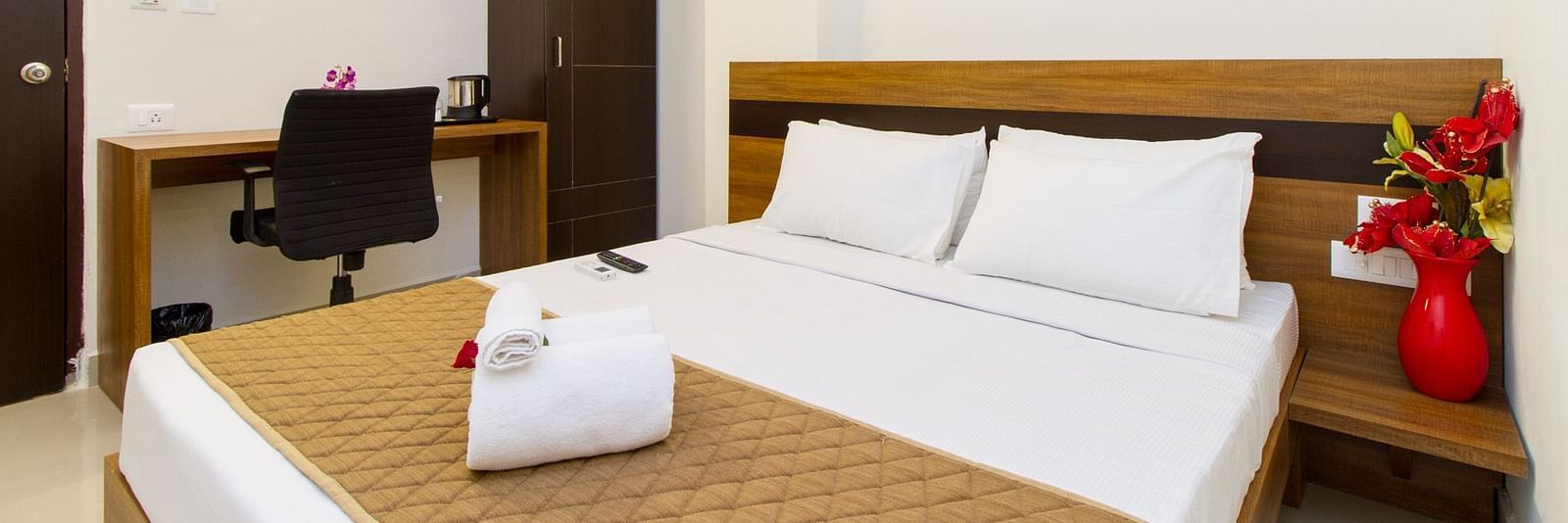 A hotel room including a double bed with brown bedding, a dark wardrobe, and a work desk and chair at Sanctum Suites Whitefield, Bangalore.
