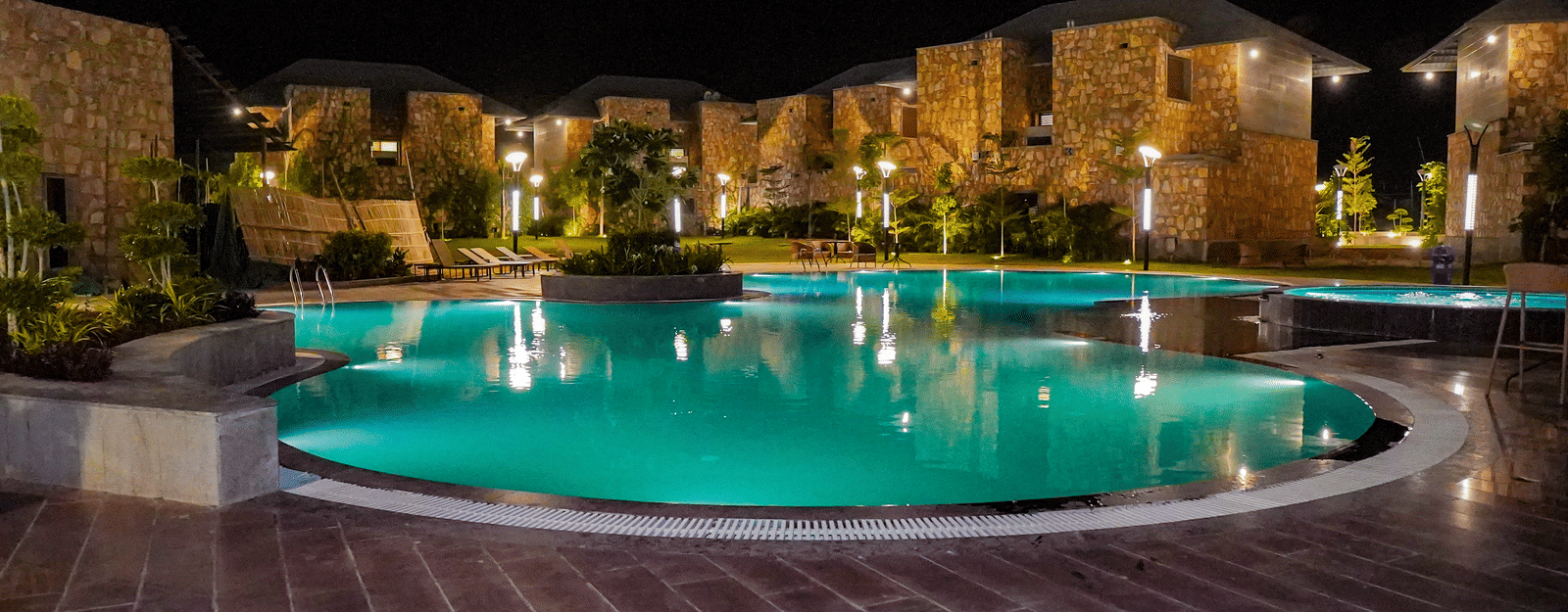 An illuminated swimming pool glows green and reflects the resort buildings at night. The surrounding area is dark with a view of the mountains.