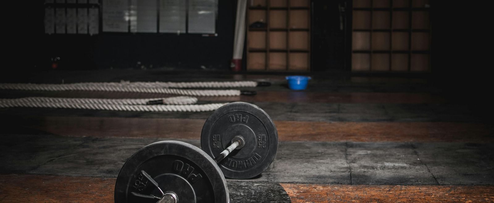A barbell with weights lies on a worn gym floor, with long metal bars in the background.