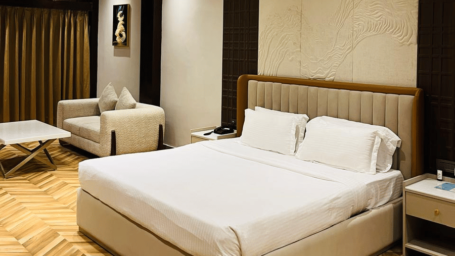 A modern hotel room with a king size bed, beige sofa, white coffee table, wooden herringbone flooring, warm lighting, and a gold and blue abstract artwork on the wall - Silver Sand Beach Resort, Havelock