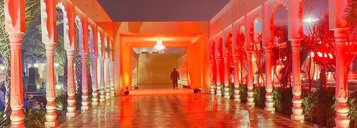 A long, arched walkway within a palace or courtyard, illuminated with a vibrant red-orange light that reflects strongly off the checkered floor tiles at night.