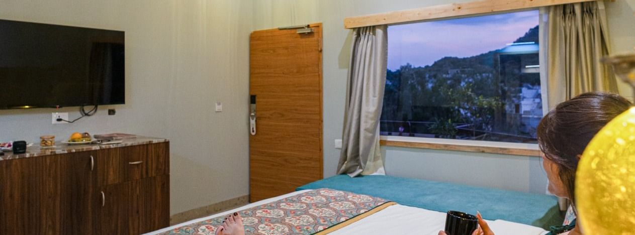 Guest relaxing on bed at Bamboo Saa Resort & Spa, Udaipur with hills view from window.