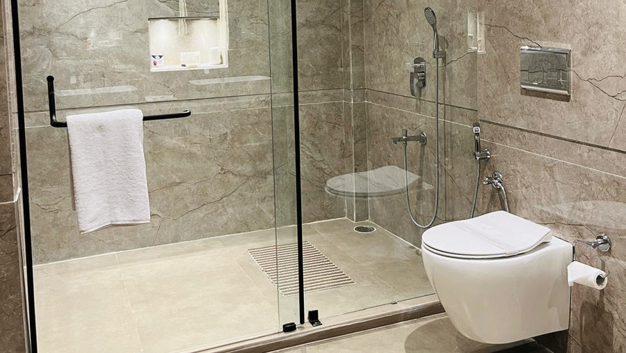 A modern hotel bathroom featuring beige marble walls and flooring, a glass-enclosed shower area with a rainfall showerhead and handheld shower, and a wall-mounted toilet with sleek fixtures under recessed ceiling lights - Silver Sand Beach Resort, Havelock