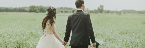 A bride and groom in wedding attires walking thorugh a field while holding hands