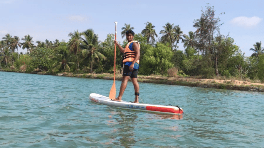 One person standing on paddle-boards in the river under a bright blue sky, holding paddles at Paradise Lagoon Resort, Udupi.