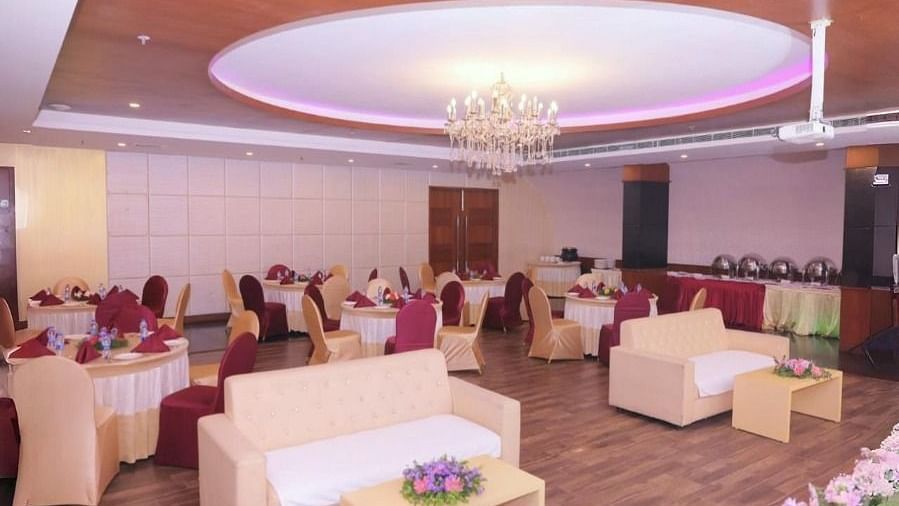 A banquet hall in Dhanalakshmi Srinivasan Hotels, Perambalur, with round tables, chairs, sofas, and a floral arrangement in the foreground. The hall has wooden flooring and a chandelier.
