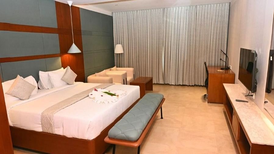 A hotel room at Dhanalakshmi Srinivasan Hotels, Perambalur, featuring a double bed, two benches, a TV on a console, a desk with a chair, two armchairs, and closed curtains.