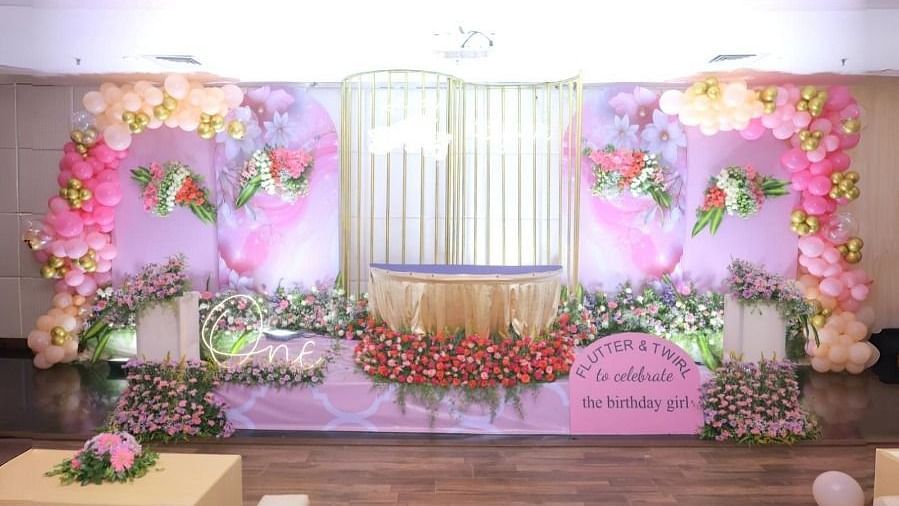 A decorated hall in Dhanalakshmi Srinivasan Hotels, Perambalur, set up for a celebration with a stage adorned with pink and gold balloons, flowers, and a banner. There are sofas and a coffee table in the foreground.