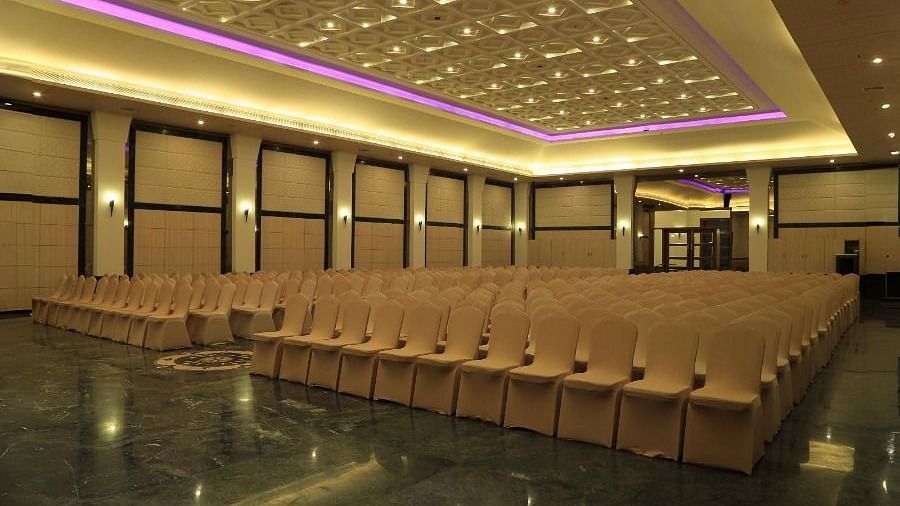 A large empty hall in Dhanalakshmi Srinivasan Hotels, Perambalur, with rows of chairs facing a stage. The hall has dark marble flooring and a patterned ceiling with recessed lighting.