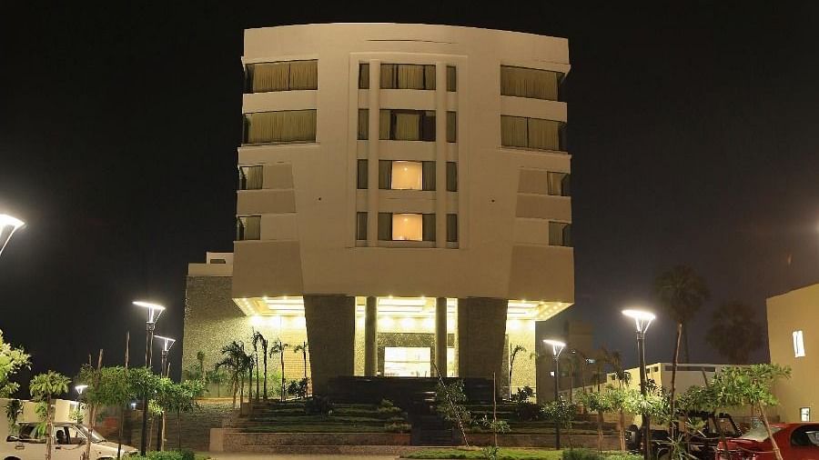 The exterior of Dhanalakshmi Srinivasan Hotels, Perambalur, a multi-story building with light-coloured facades and yellow-tinted windows, seen from a paved road under a blue sky.