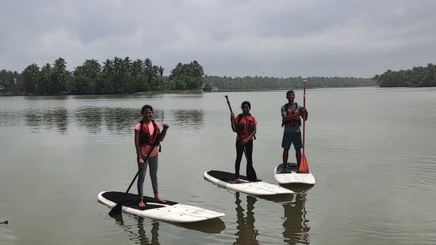 Three people balancing on paddle-boards on a cloudy day with calm waters and distant treeline at Paradise Lagoon Resort, Udupi.