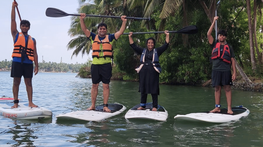 Three people standing on paddle-boards in the river near palm trees, balancing with paddles in hand at Paradise Lagoon Resort, Udupi.