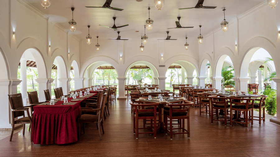 A spacious restaurant interior with rows of wooden dining tables, high ceilings with fans, and large arched openings | Le Pondy