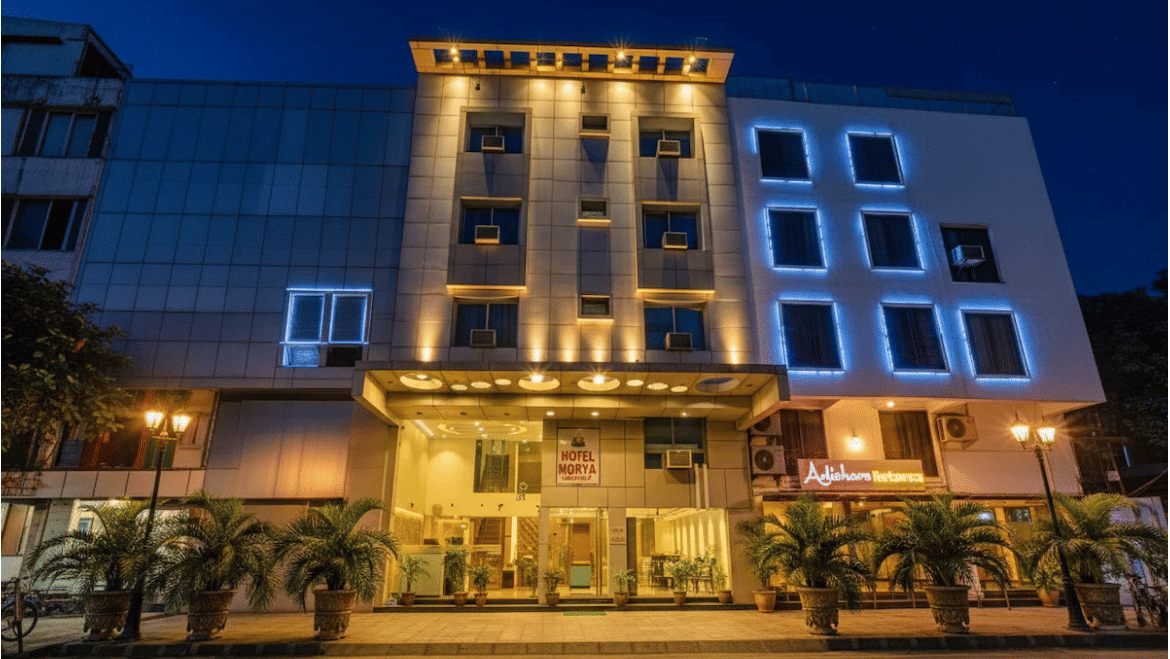 Front view of Hotel Maurya, Bhopal at night, multi-storey building with lit windows, glass entrance, and potted palms along the driveway.
