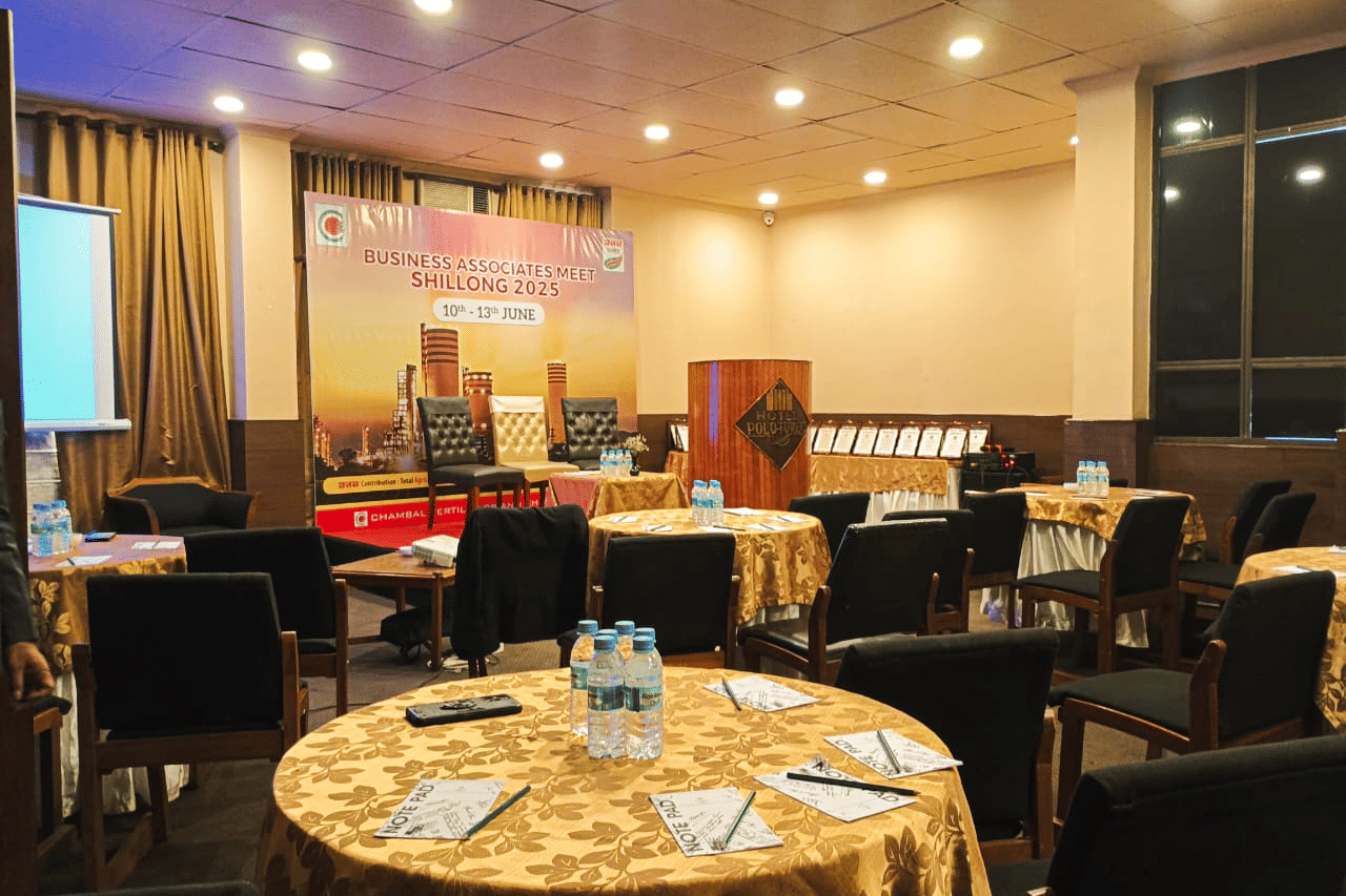A conference room at Polo Hotel Shillong set up for a meeting, with a stage, podium, and round tables with chairs.