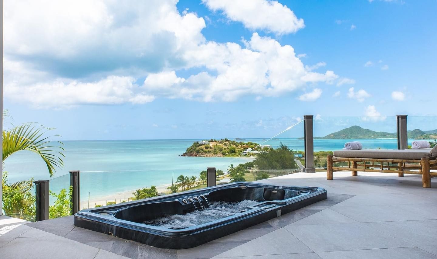  A stunning outdoor jacuzzi at Tamarind Hills Resort and Villas - Antigua, offering panoramic views of a turquoise ocean and a clear blue sky with scattered clouds. The jacuzzi is black and appears to be built into a light-colored patio.