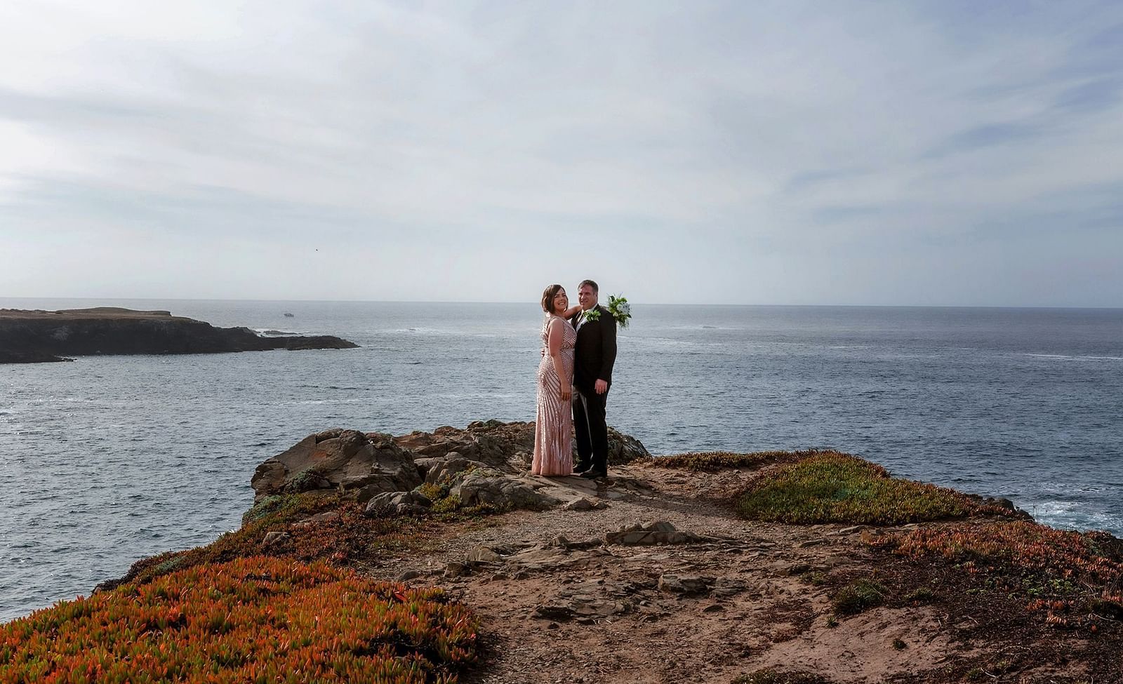Bride and groom embracing on a rocky cliff with the vast ocean behind them