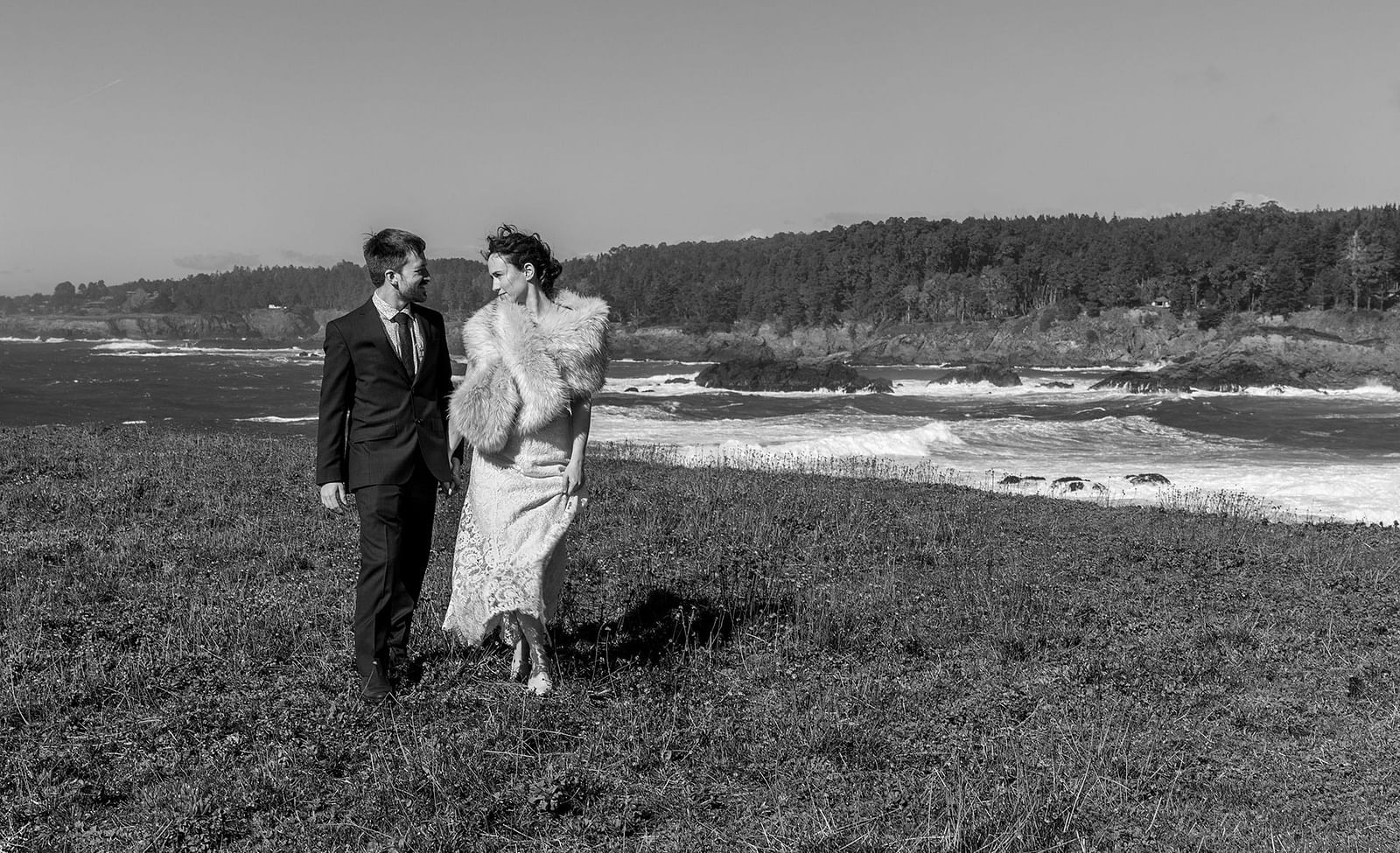 A black-and-white image of a couple walking near the ocean on a breezy day