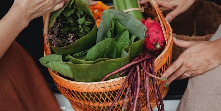 A pair of hands holds a woven basket overflowing with fresh green leafy vegetables and red-purple beans, some wrapped in large leaves.
