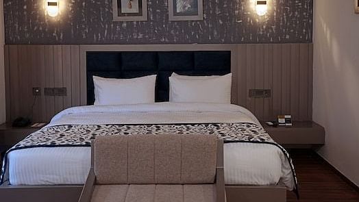 A hotel bedroom at Grand Continent, Anjuna featuring a large bed, a small sofa at the foot, a decorative textured wall behind the bed with two framed pictures and two sconce lights.