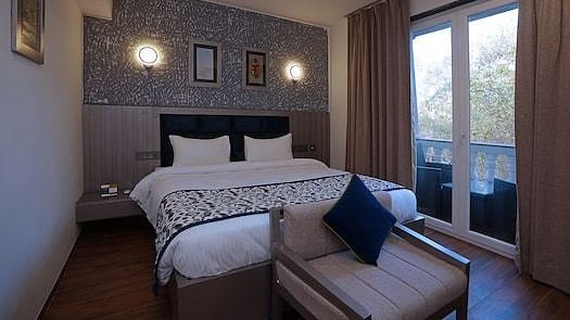 A hotel bedroom at Grand Continent, Anjuna featuring a large bed, a sofa at the foot, and a balcony with open curtains visible on the right. The wall behind the bed has decorative textured panelling and sconce lights.
