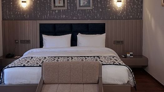 A hotel bedroom at Grand Continent, Anjuna featuring a large bed, a small sofa at the foot, a decorative textured wall behind the bed with two framed pictures and two sconce lights.